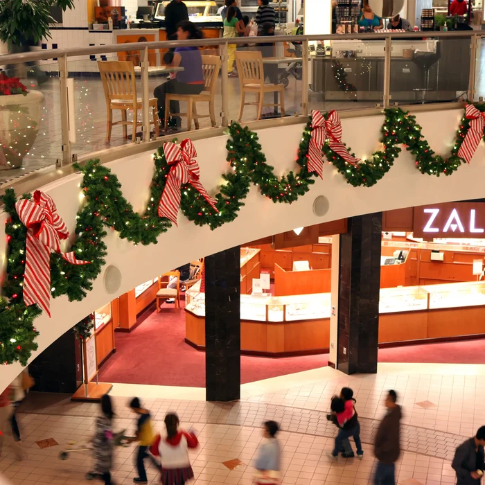 A busy indoor shopping mall scene with holiday green garlands and large red-and-white striped bows draped along a curved upper walkway. People are visible walking on the lower floor, and retail counters and a ZAL sign are visible in the background.