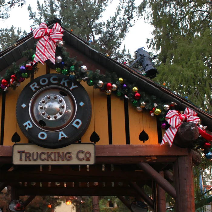 A rustic wooden sign reading 'Rocky Road Trucking Co' mounted on a decorated building with Christmas garlands, colorful ornaments, red-and-white striped bows, and evergreen trees in the background.