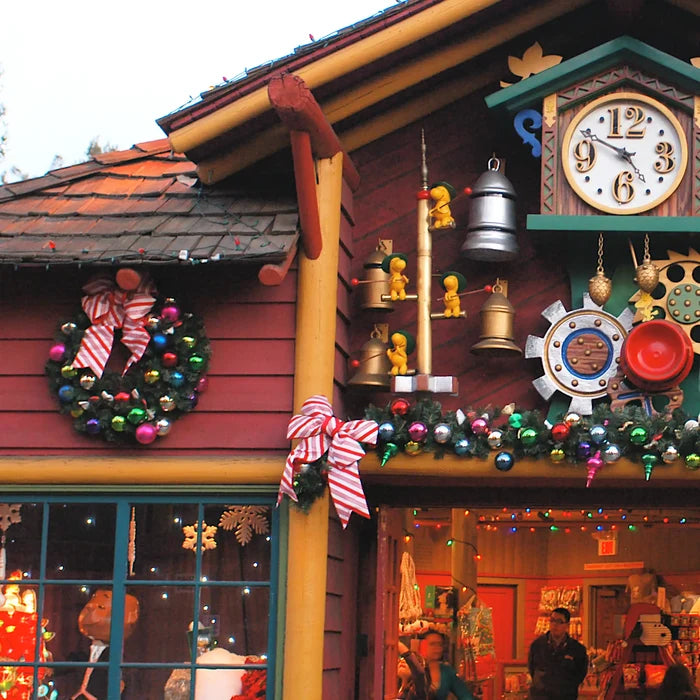 A festive holiday storefront facade with a large red Christmas wreath, colorful ornaments, a clock, hanging bells, and garlands. A real outdoor environment and storefront interior are visible.