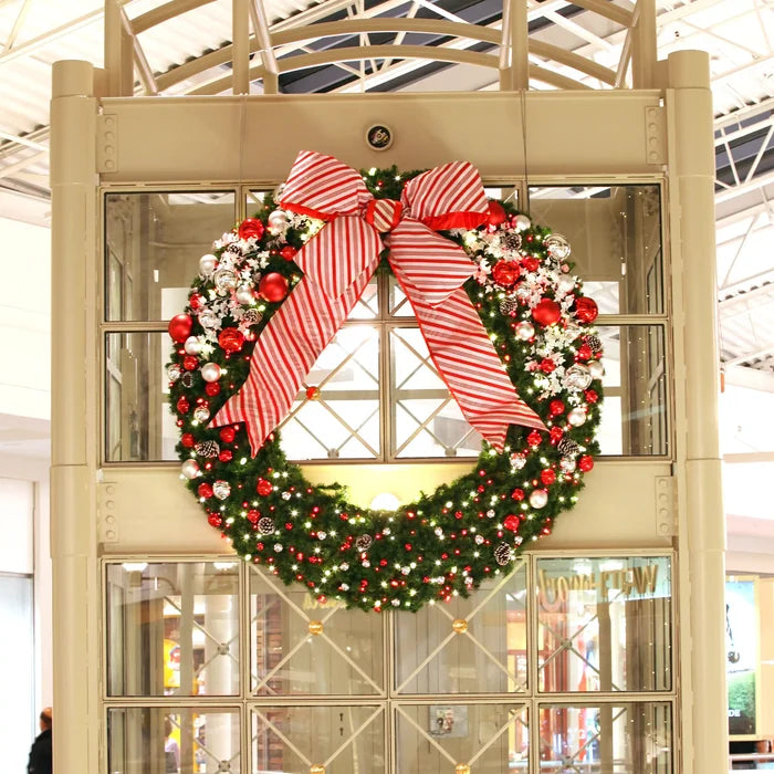 A festive Christmas wreath hanging on a large cream-colored glass display case in a shopping mall. The wreath is decorated with green pine branches, red ornaments, white berries, and a large red and white striped bow on top.