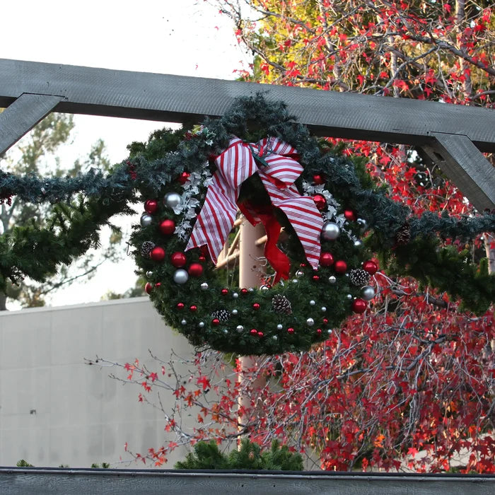 A festive Christmas wreath hanging from a wooden trellis, decorated with a large red and white striped bow, green pine branches, red and silver ornaments, and a small wooden star.