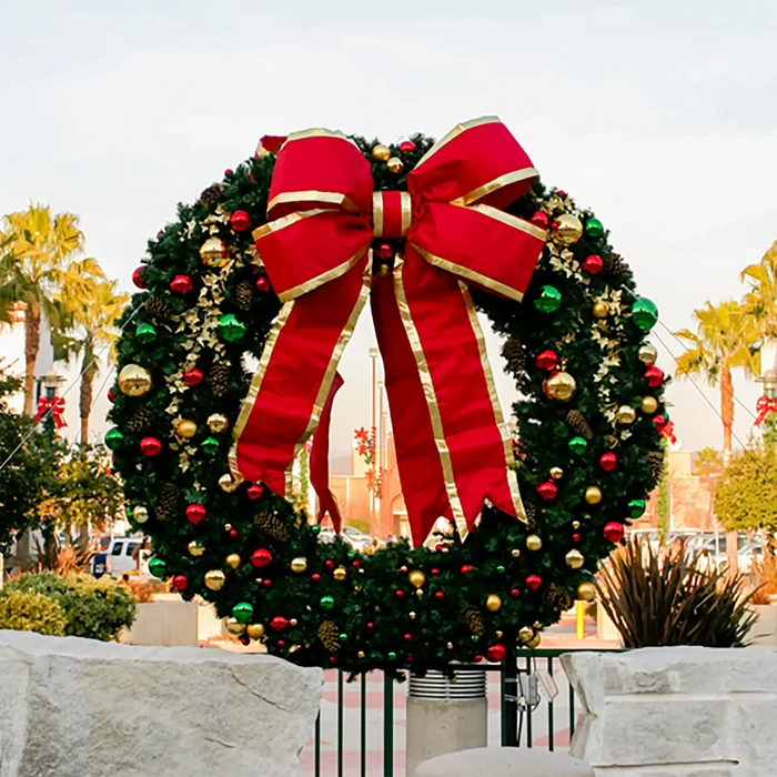 A large Christmas wreath with a giant red bow and gold trim, decorated with red, green, and gold ornaments, set against an outdoor background with palm trees and a stone wall.