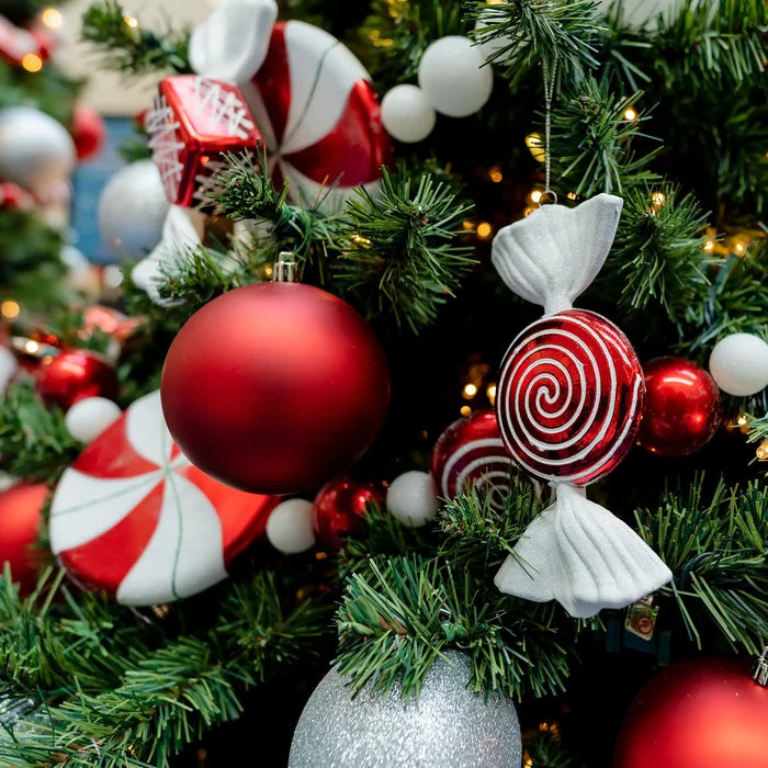 Close-up of a decorated Christmas tree with red and white candy ornaments, red and white baubles, white pom-poms, and warm string lights among green pine branches.
