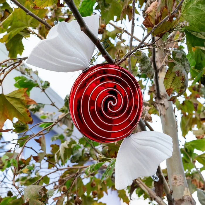 A red spiral-patterned hanging ornament or decorative piece suspended from a tree branch, with two white petal-like wings or panels attached on either side, photographed outdoors among green and brown leaves.