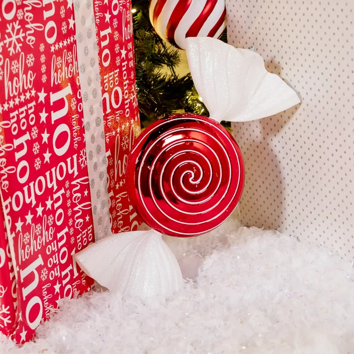 A festive holiday-themed product display featuring a glossy red spiral lollipop ornament, red and white patterned gift wrapping with 'ho ho ho' text, white marshmallows, and a red-and-white striped candy cane against a light beige dotted backdrop.
