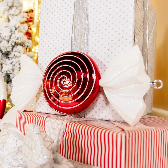 A red spiral lollipop Christmas ornament with white bow-like wings, displayed on top of wrapped gift boxes with silver and red striped paper.