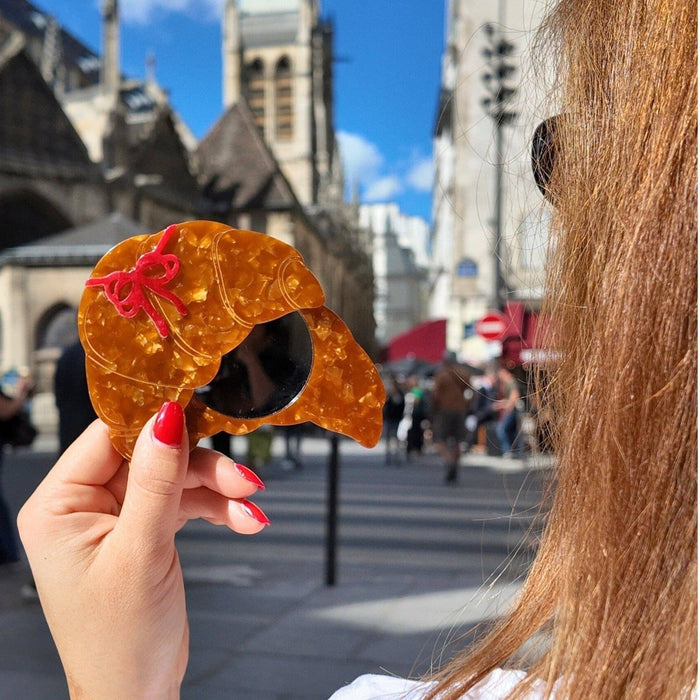 Hand-painted bow croissant compact mirror held up in Parisian street with historic architecture backdrop