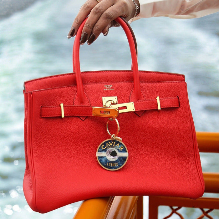 Red Hermès Birkin bag with hand-painted caviar luxury charm keychain held by model near water